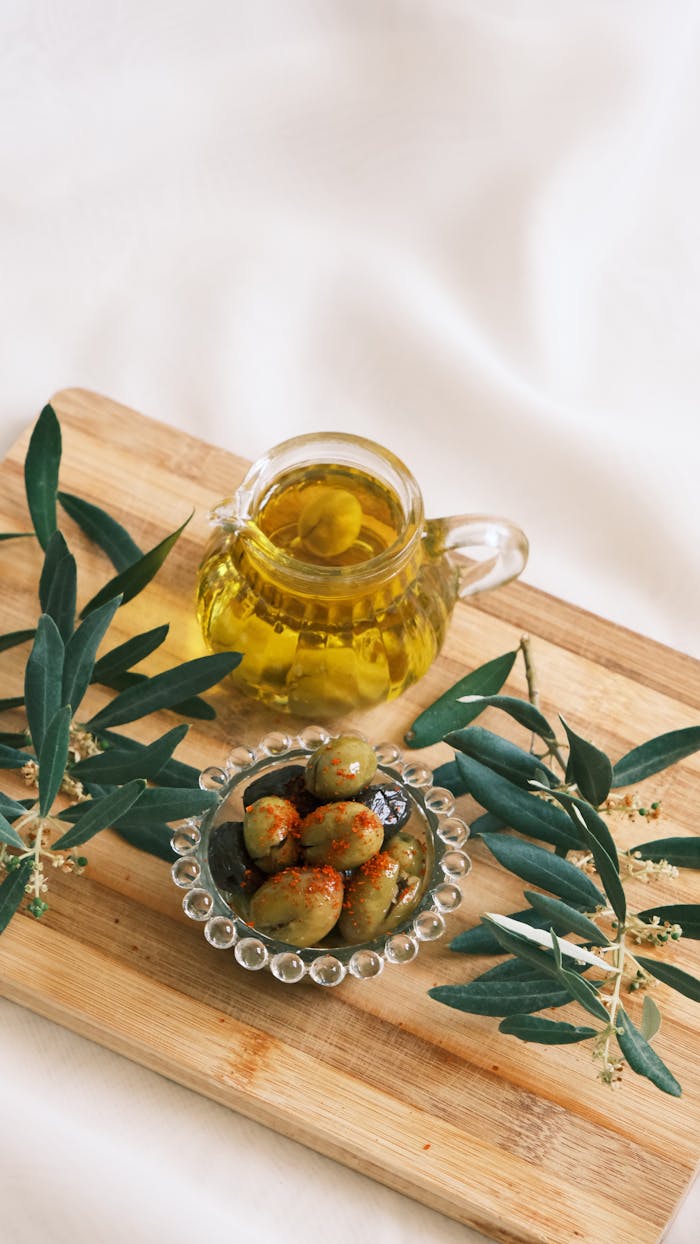 who-we-are Glass jug of olive oil and a bowl of olives on a wooden cutting board with olive branches.