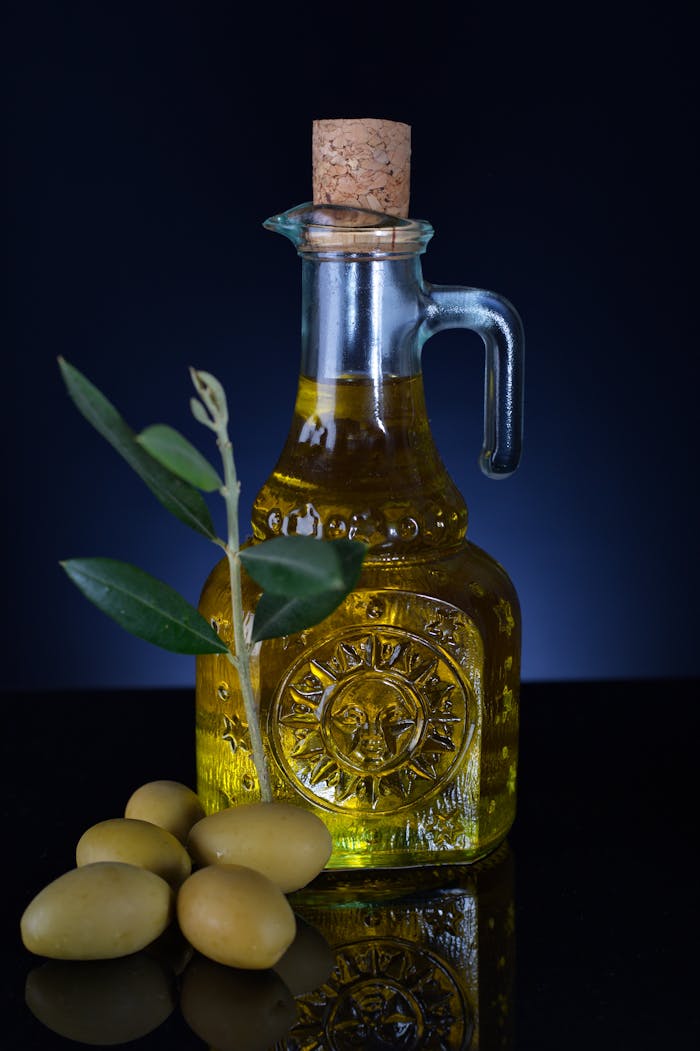 Close-up of a decorative glass jar filled with olive oil, accompanied by olives and leaves, on a reflective surface.